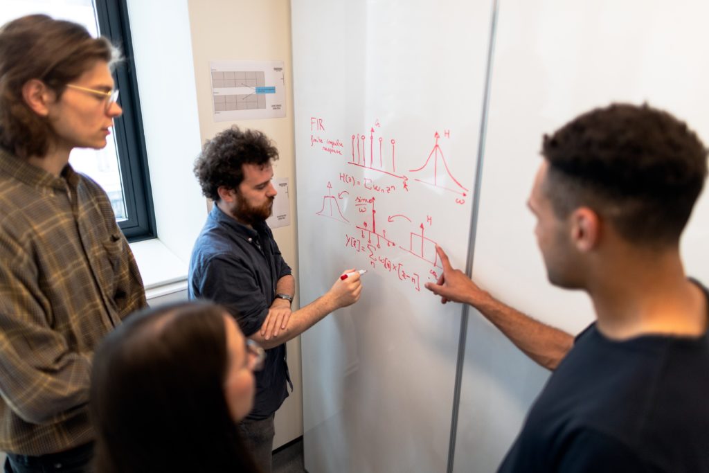 A picture of a team of 4 people standing around a white board. 2 people are pointing at the board, 1 with a red marker doing calculations, while the other 2 stand and look onward. Much like these students, article is about the best data science courses and top online programs.